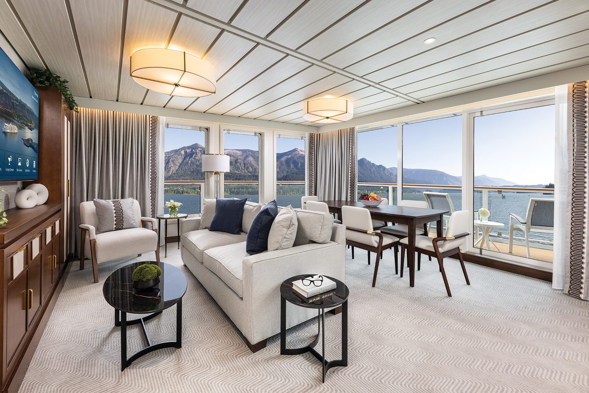 A Signature Suite living room aboard American Encore with a sofa, dining table for four and wraparound windows facing the mountains