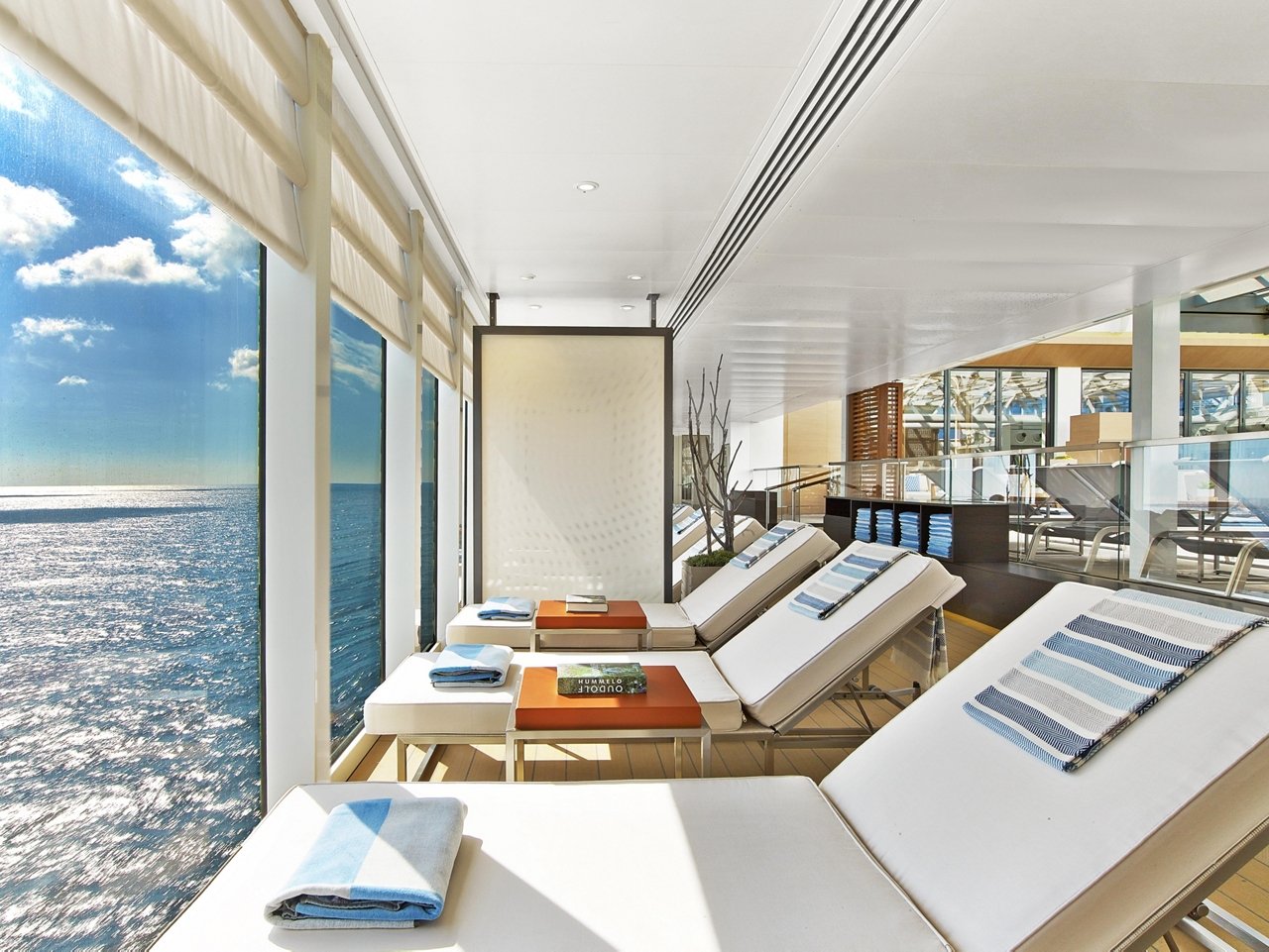 White loungers with striped towels line the main pool deck of a Viking ocean ship, beside floor-to-ceiling windows looking out to sea