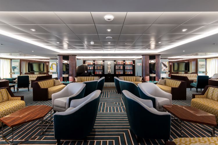 A stylish lounge with plush armchairs, patterned carpet and a backlit library wall aboard an Atlas Ocean Voyages ship