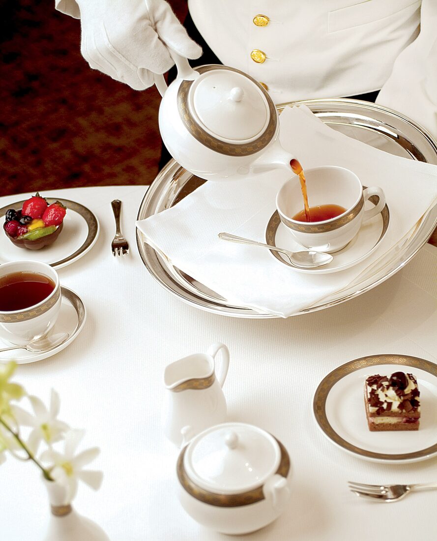 A white-gloved Cunard waiter pours tea from a china teapot onto a silver tray set with pastries and fresh fruit tarts