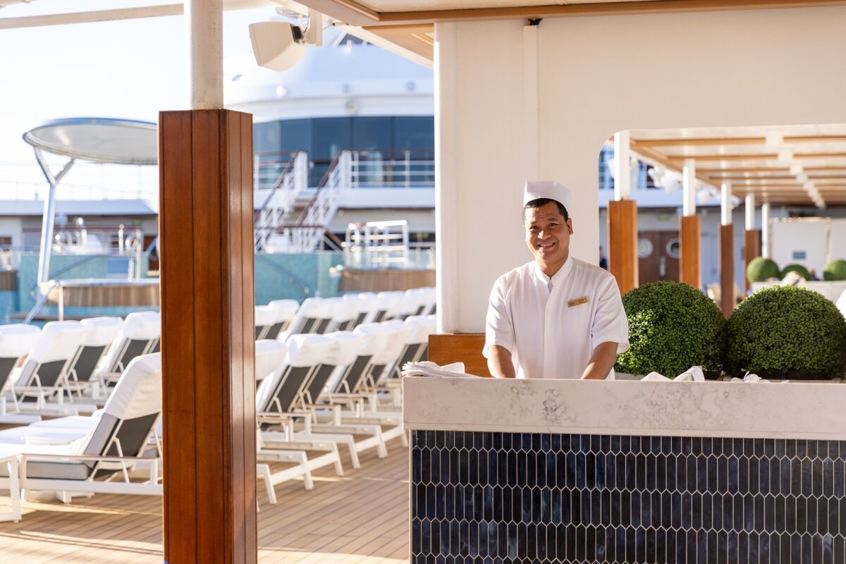 A smiling crew member in whites stands at a marble-topped pool deck bar aboard a Regent Seven Seas ship