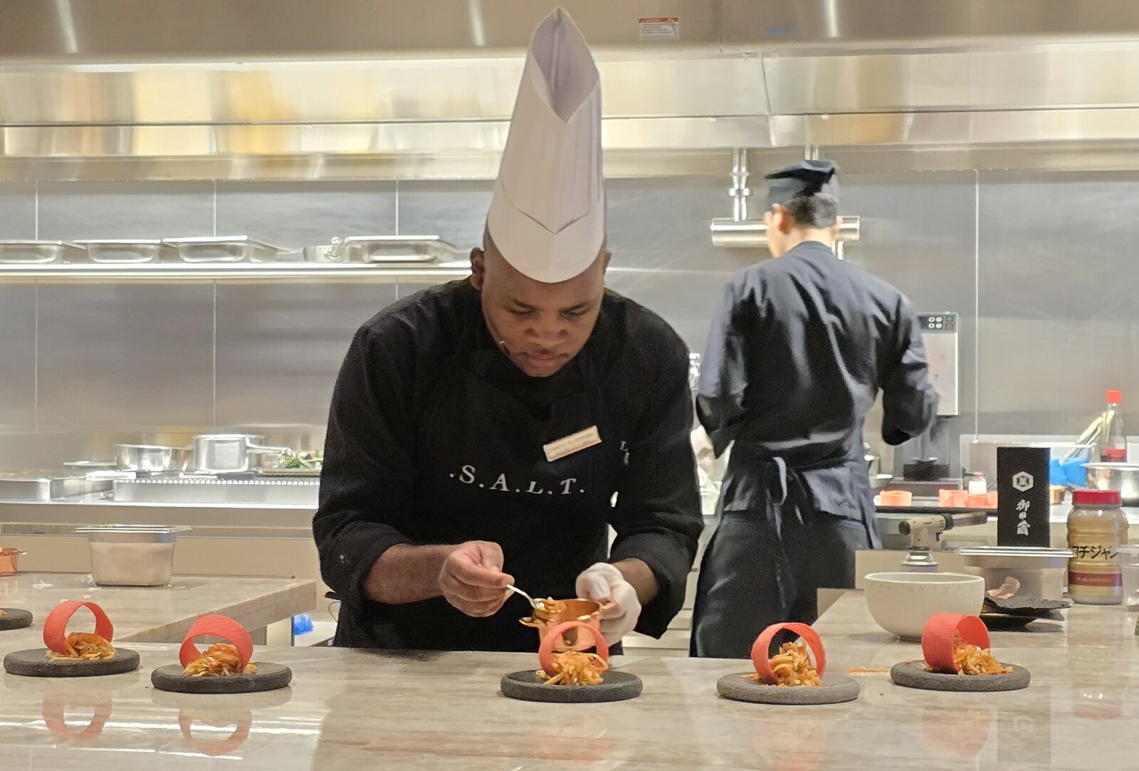 A chef in a S.A.L.T. apron carefully plates a row of artfully presented dishes at a live cooking counter aboard a Silversea ship